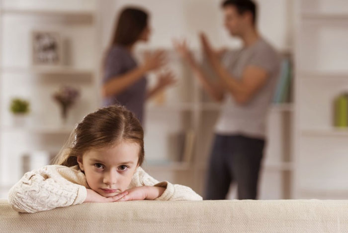 An upset girl on the couch and her parents are fighting in the background.